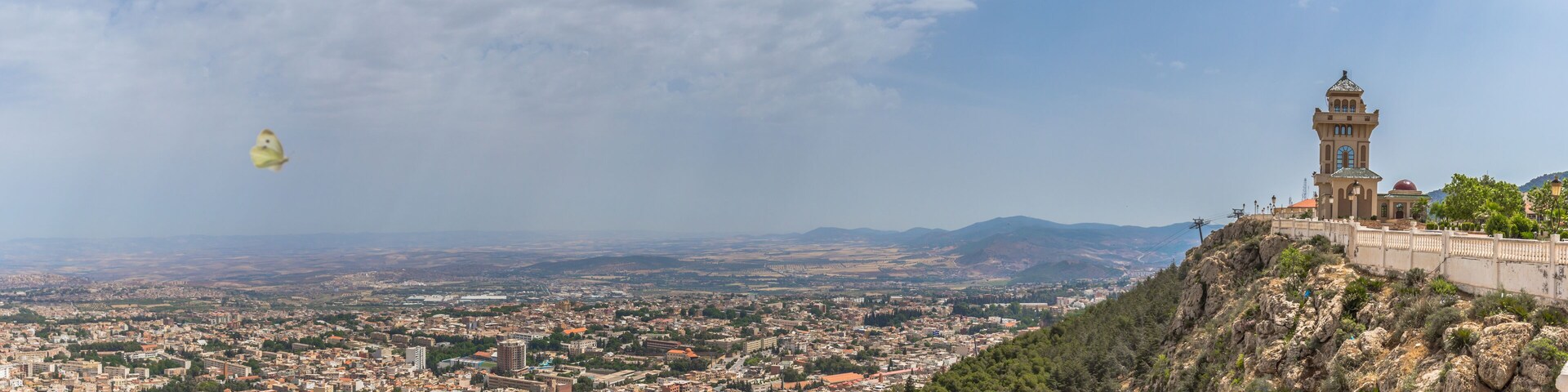 Un paysage urbain avec une tour imposante sur une colline, une vallée en contrebas et un cerf-volant qui vole dans le ciel.