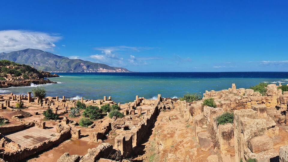 Ancient ruins by a coastal cliff with a mountain in the background.