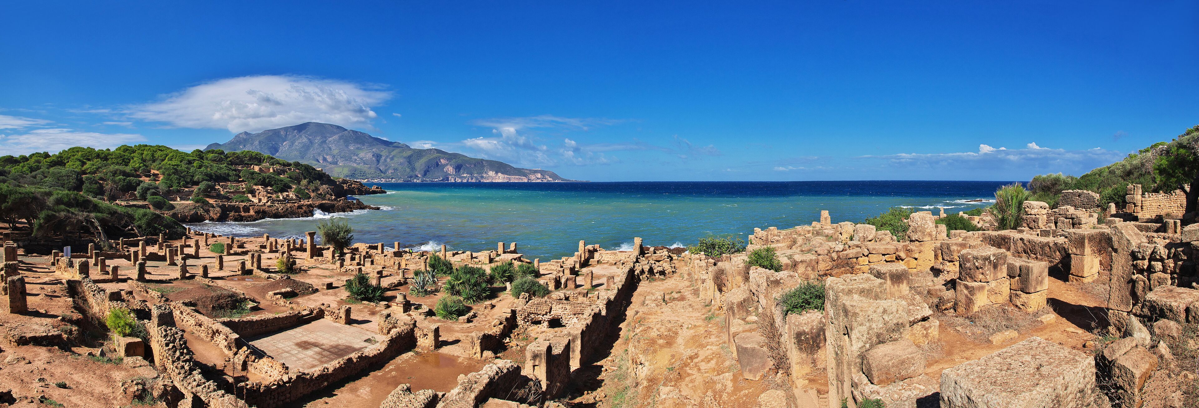 Ancient ruins by a coastal cliff with a mountain in the background.
