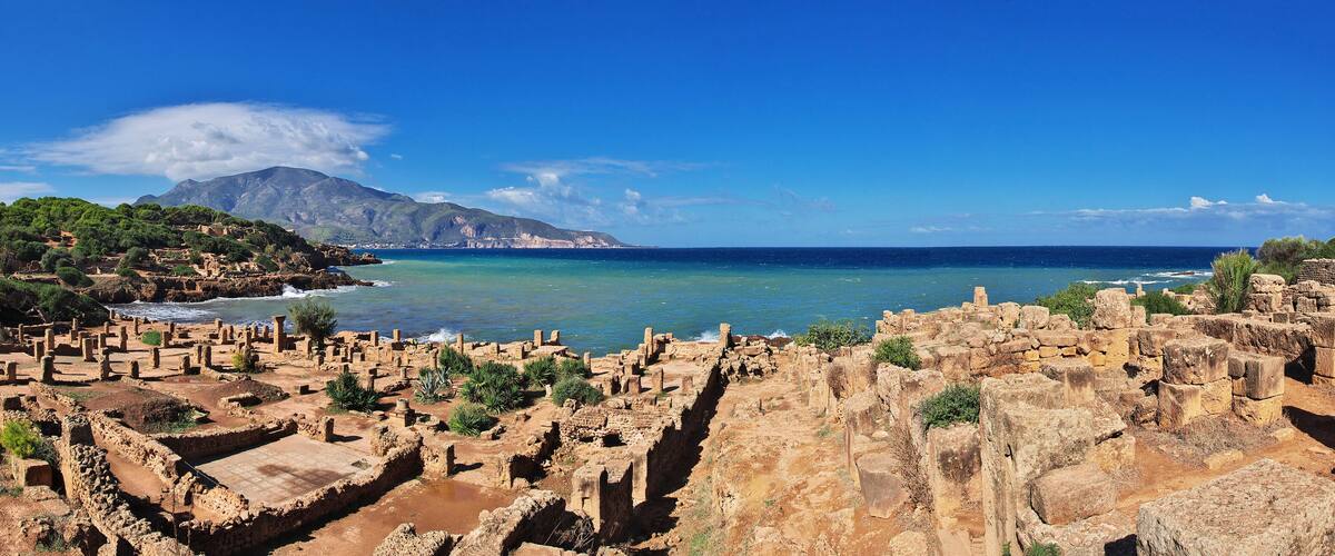 Ancient ruins by a coastal cliff with a mountain in the background.