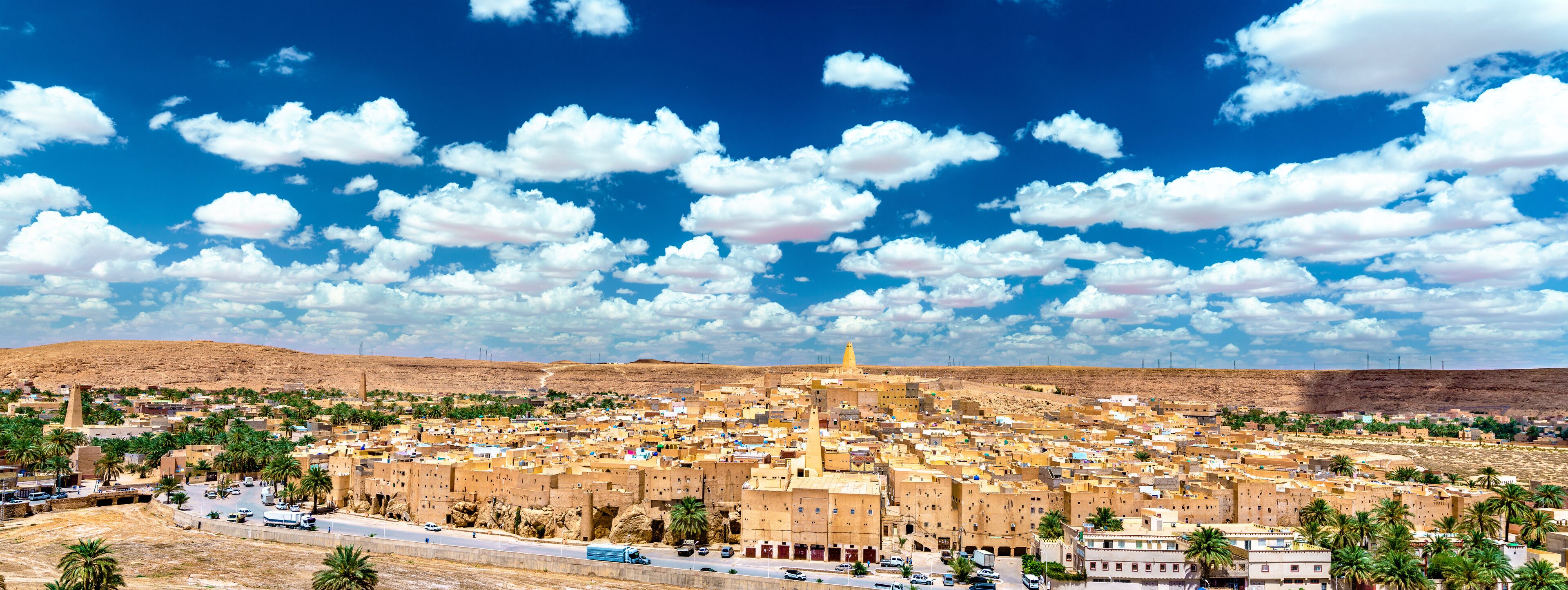 A cityscape with a prominent mosque, palm trees, and a clear blue sky with scattered clouds.