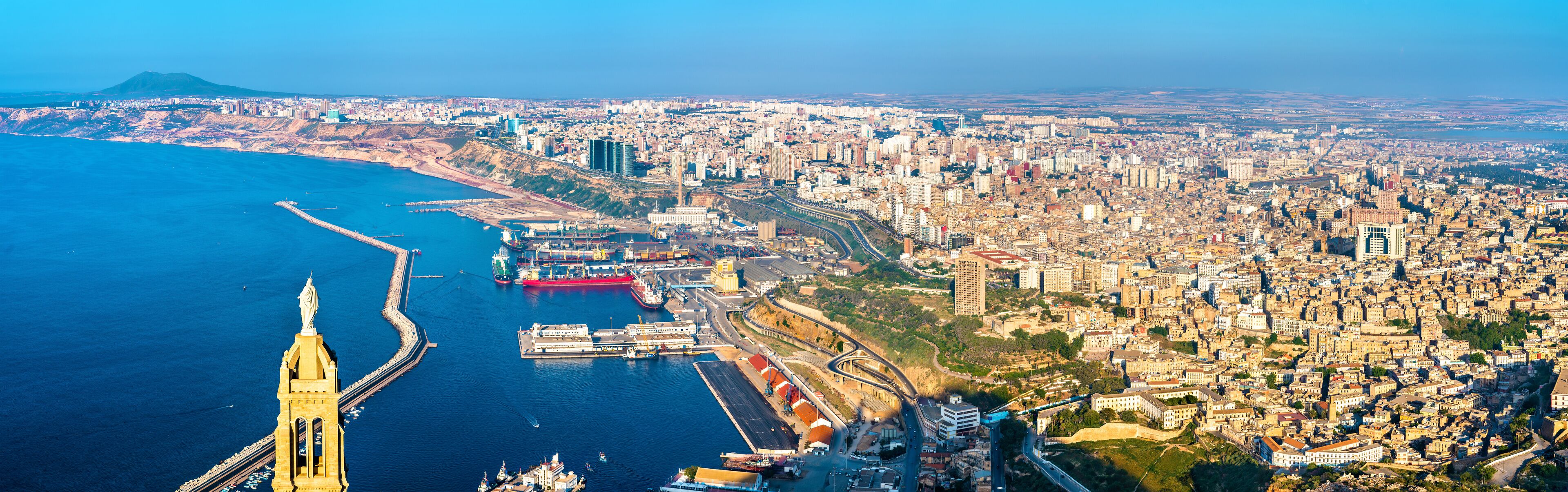 An aerial view of a coastal city with a prominent tower, a harbor with ships, and a dense urban area.