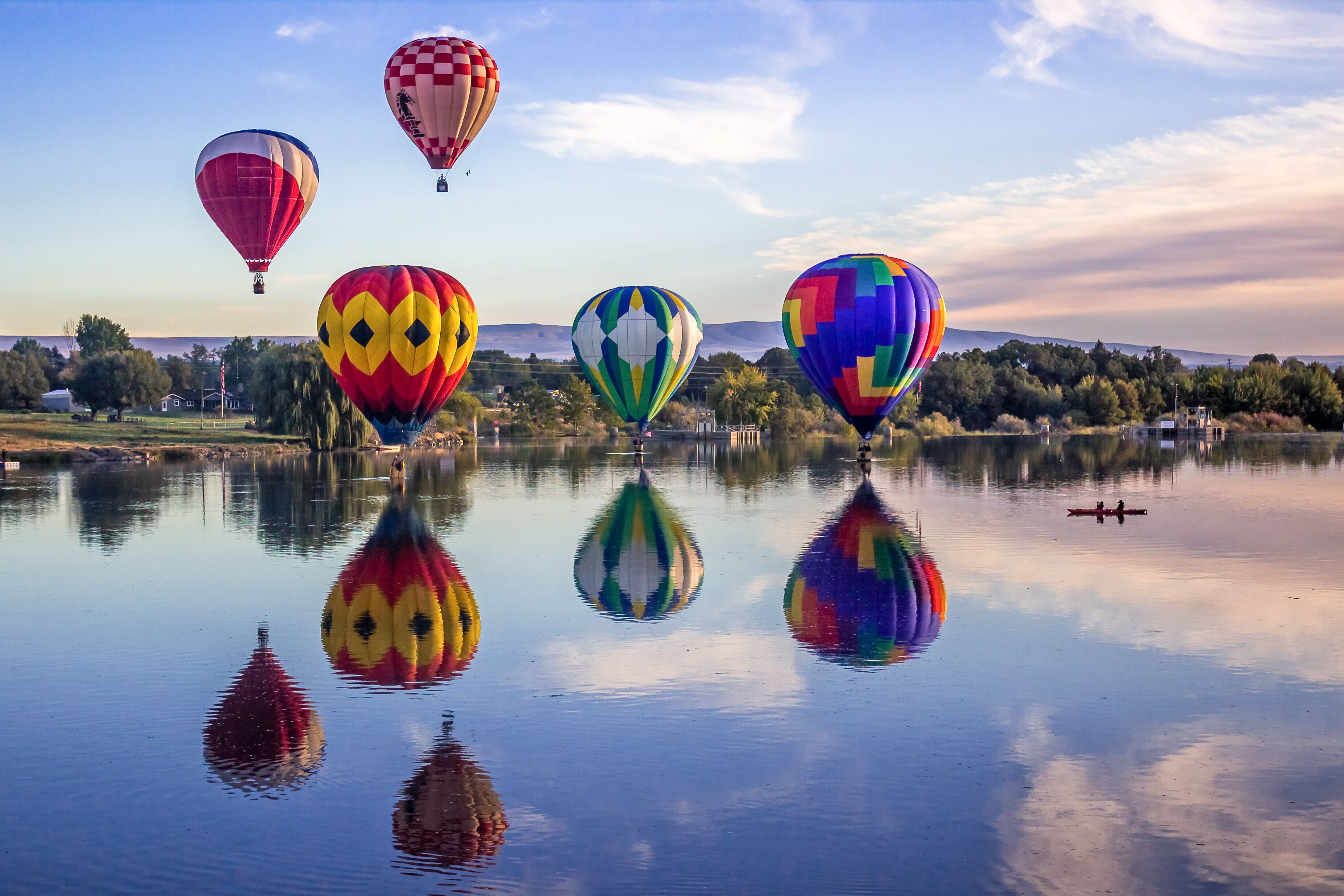 Hot air balloons over a calm lake with reflections, surrounded by trees and a distant mountain range.