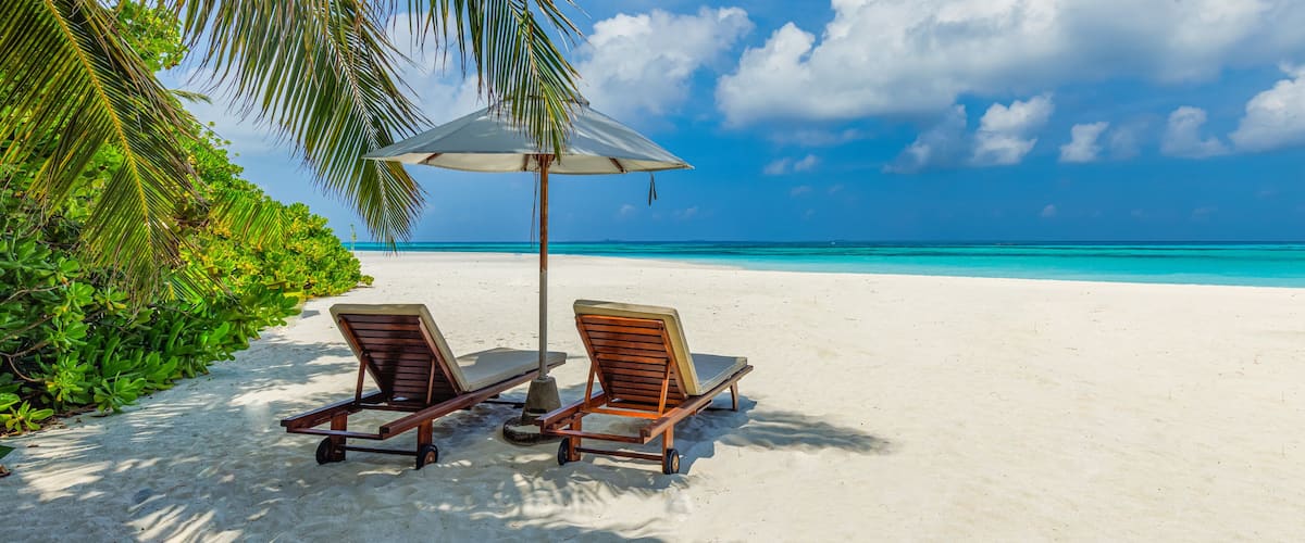 Two wooden lounge chairs under a beach umbrella on a sandy beach with clear turquoise water and palm trees.