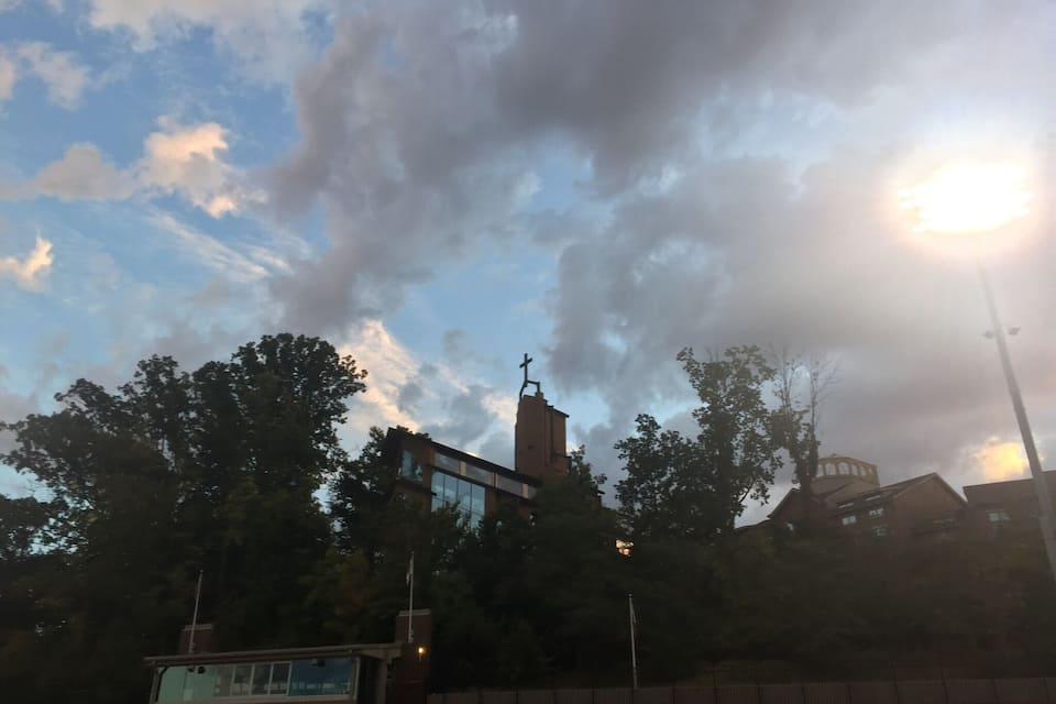 Een stadion met lege rode zitplaatsen, een hoog gebouw met een kruis bovenop en bomen onder een licht bewolkte lucht.