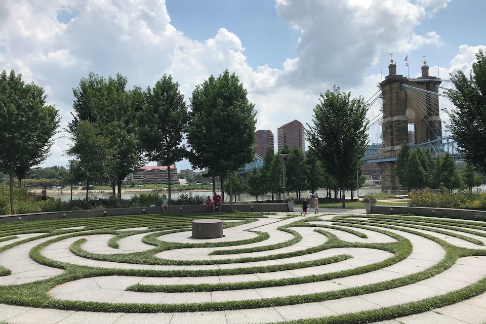 Un jardín ajardinado con un laberinto, árboles y un puente al fondo.