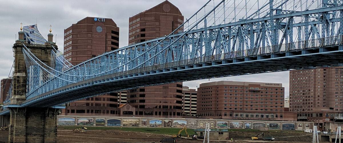 A blue suspension bridge over a river with buildings in the background.