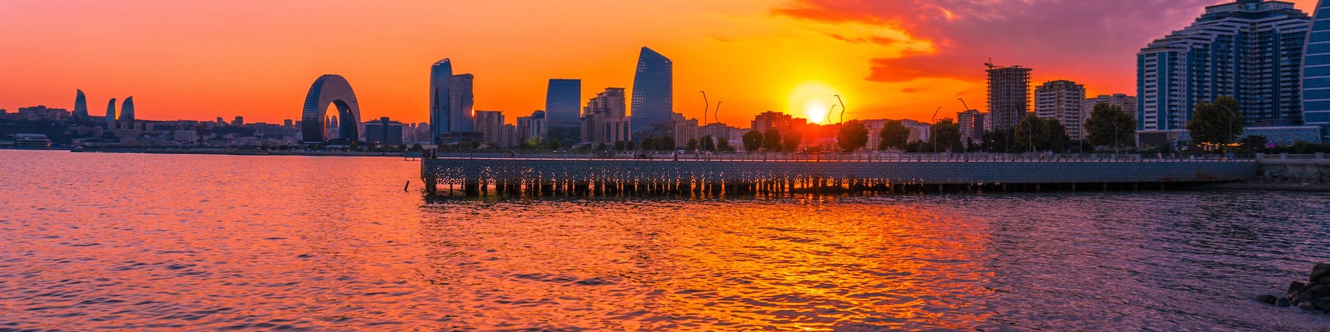 A sunset over a city skyline with a prominent arch bridge and modern buildings reflecting on the water.
