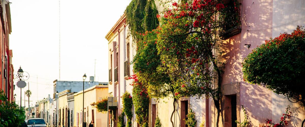 A cobblestone street lined with buildings, some with balconies and flowering plants.