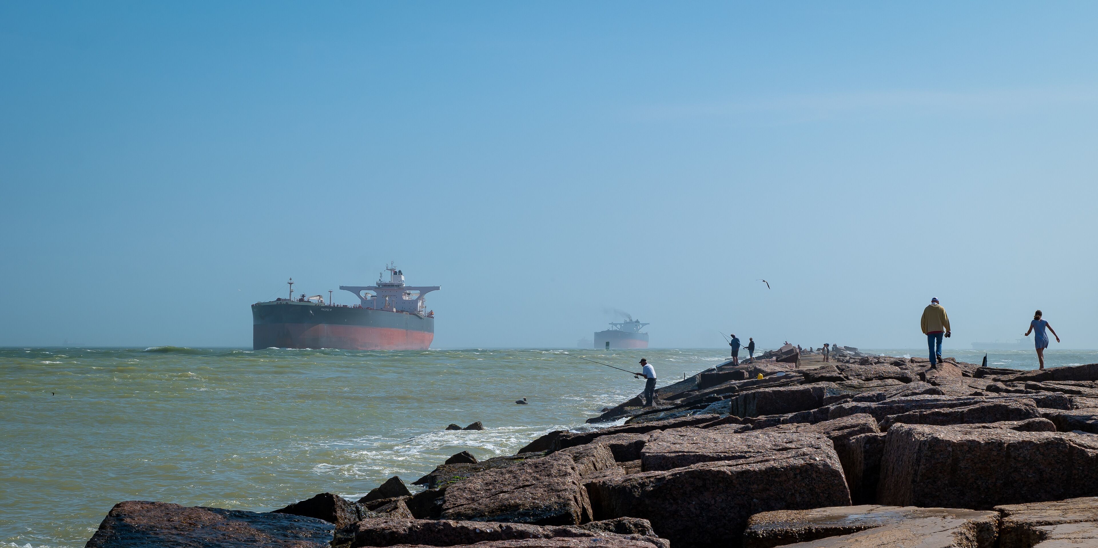 The South Jetty concrete sidewalk, with people fishing and walking, and oil tanker ships approaching from the Gulf of Mexico on the channel to Corpus Christi, Texas. 