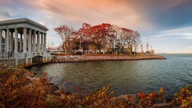 A classical building with columns, a stone bridge, a small island with red-leaved trees, and a calm body of water.