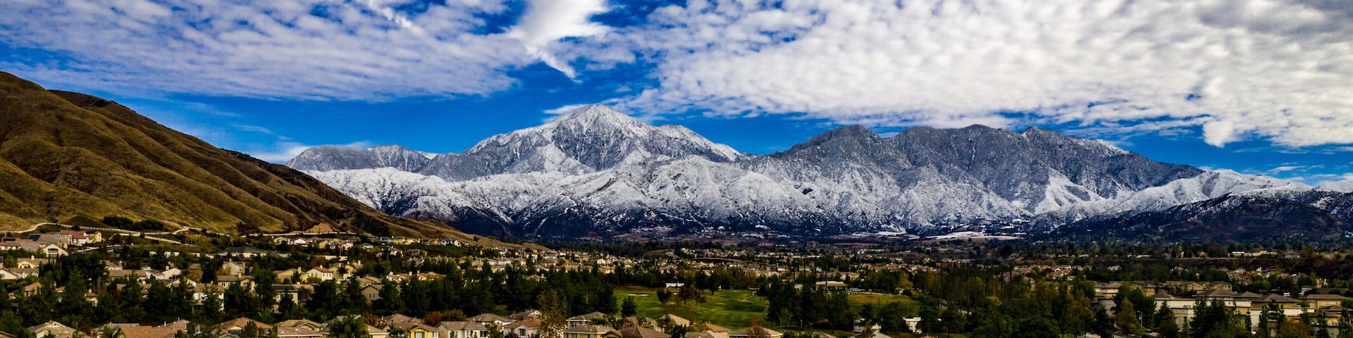 A mountain range with snow-capped peaks, a town in the foreground, and a clear blue sky with scattered clouds.