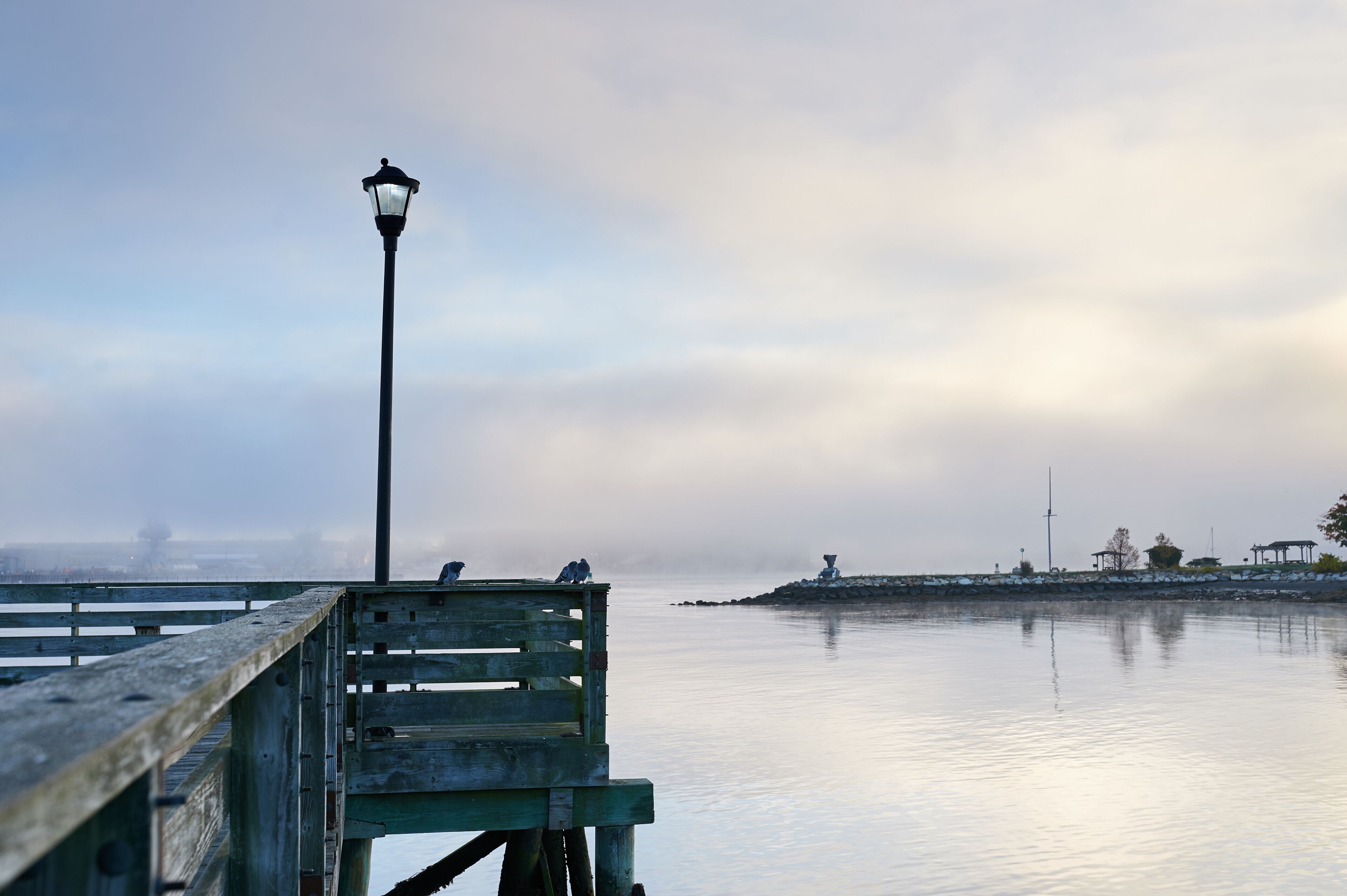 Un muelle de madera que se adentra en aguas tranquilas, un farol y un paisaje brumoso en la distancia con una pequeña isla y construcciones.