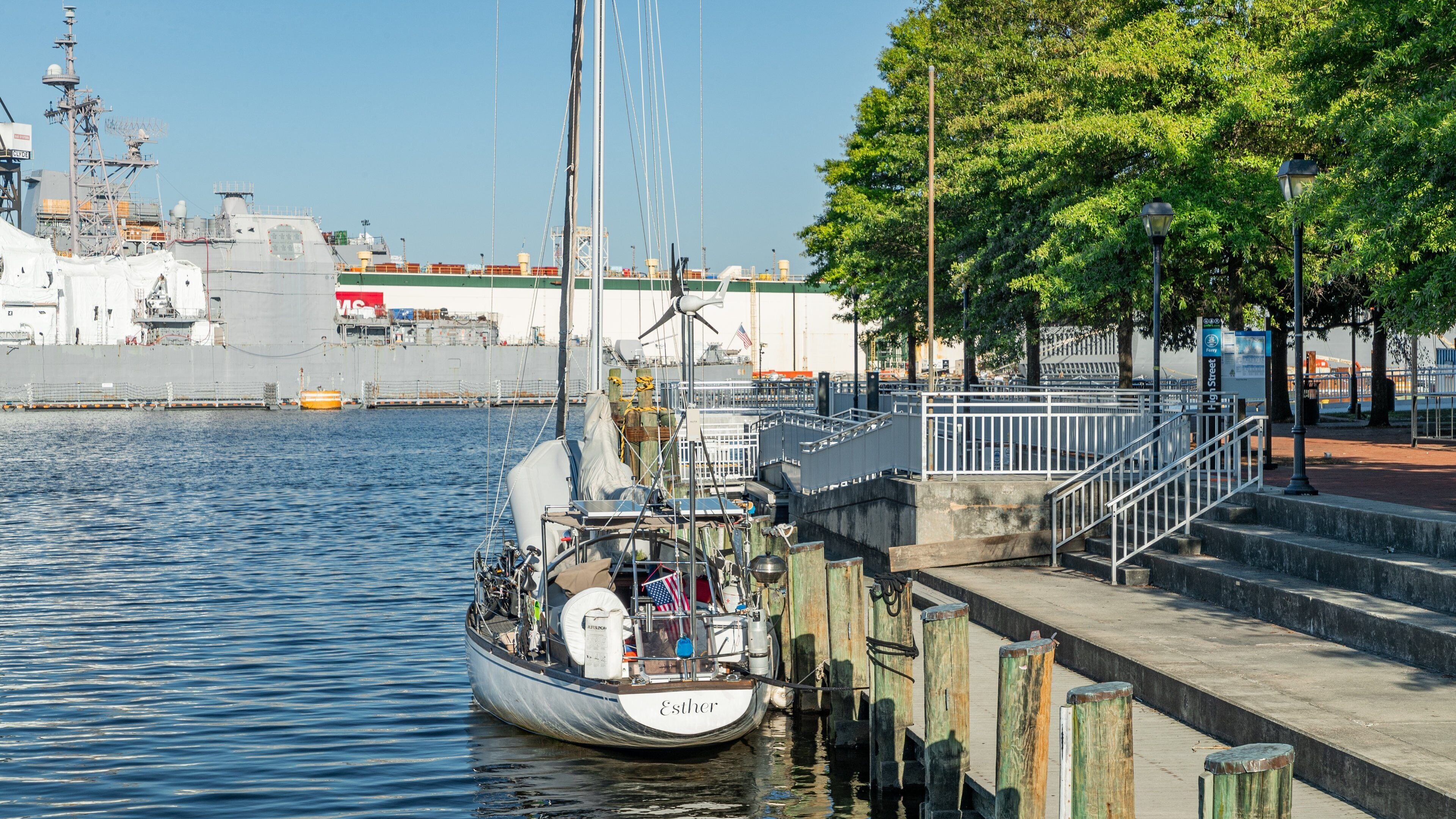 A boat named Esther docked at a pier with a large ship in the background and a park with trees and a staircase.