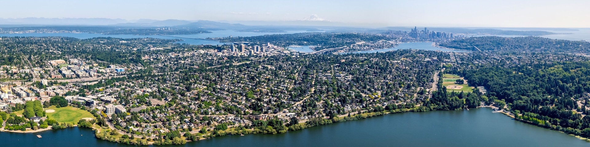 Un paysage urbain côtier avec des quartiers résidentiels densément peuplés, une vaste étendue d’eau et une chaîne de montagnes lointaine.