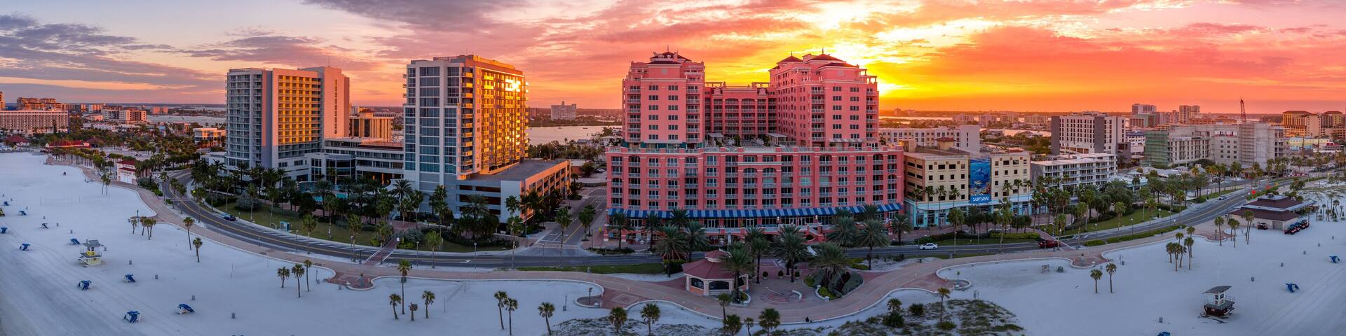 A coastal cityscape with high-rise buildings, palm trees, and a beach at sunset.