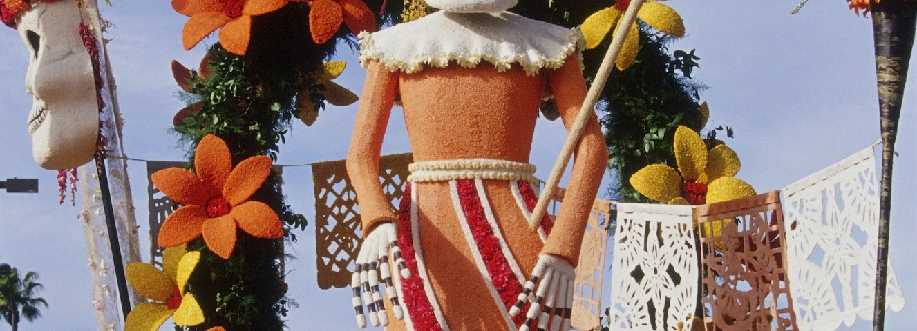 Uma figura grande e colorida de esqueleto, com coroa e guarda-sol, rodeada por flores e decorações vibrantes.