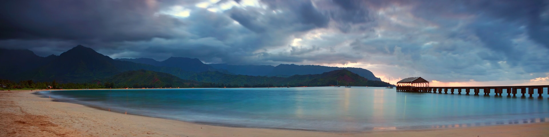 A beach with a wooden pier extending into calm waters, surrounded by mountains and a cloudy sky.
