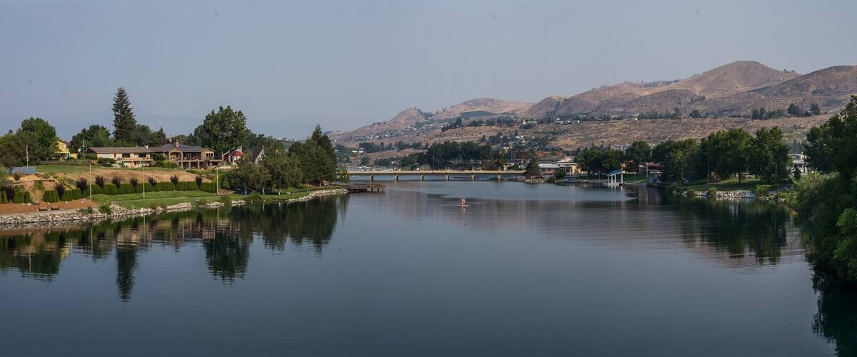 Lake Chelan panoramic landscape on a beautiful summer day, showing the lake, trees and greenery and the arid brown hills in the background.