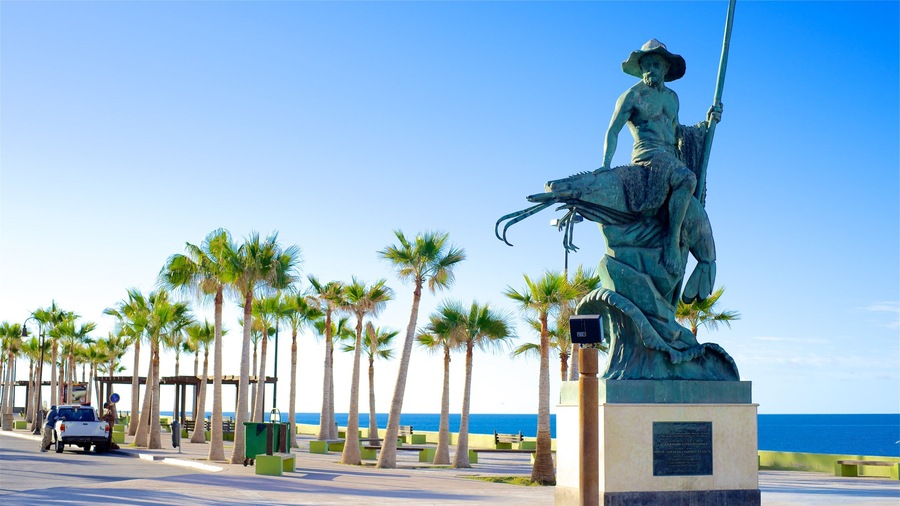Estatua de una mujer con una lanza sobre un pedestal, palmeras y una playa al fondo.