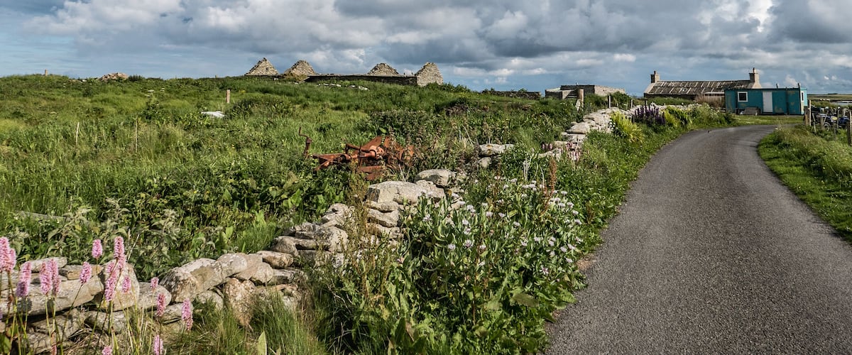 A rural landscape with a stone wall, a gravel path, and several stone structures in the distance.