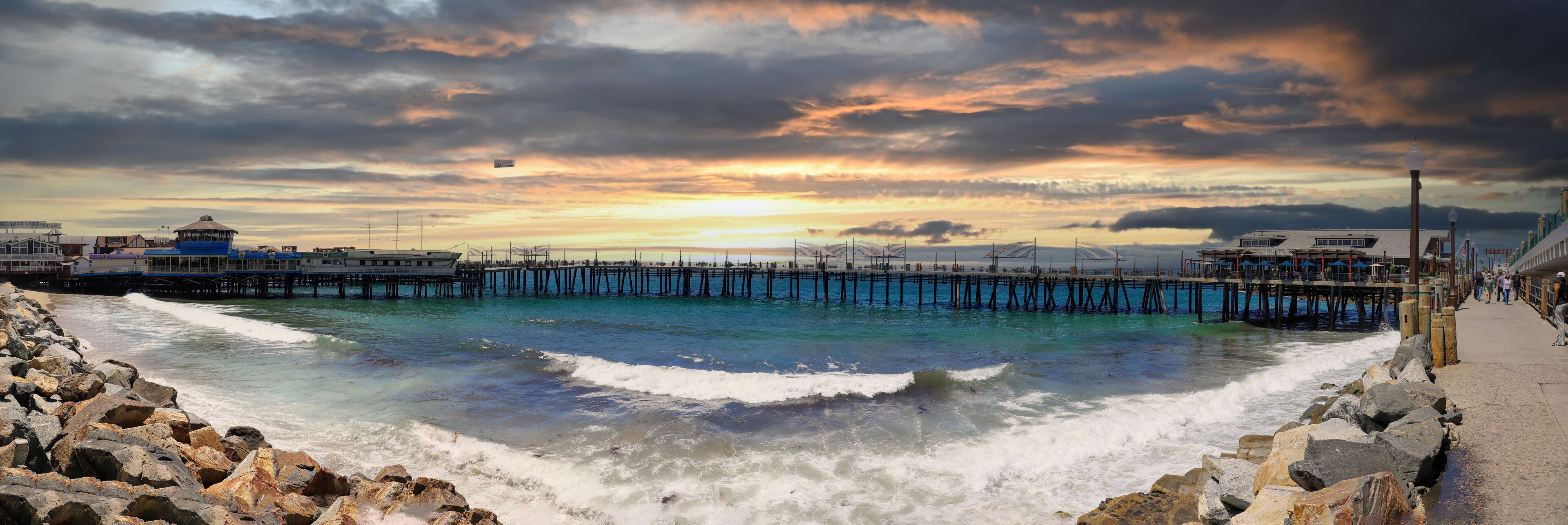 a panoramic of a gorgeous summer landscape at the Redondo Beach pier with blue ocean water and waves rolling into the brown sand on the shore with large rocks with powerful clouds at sunset