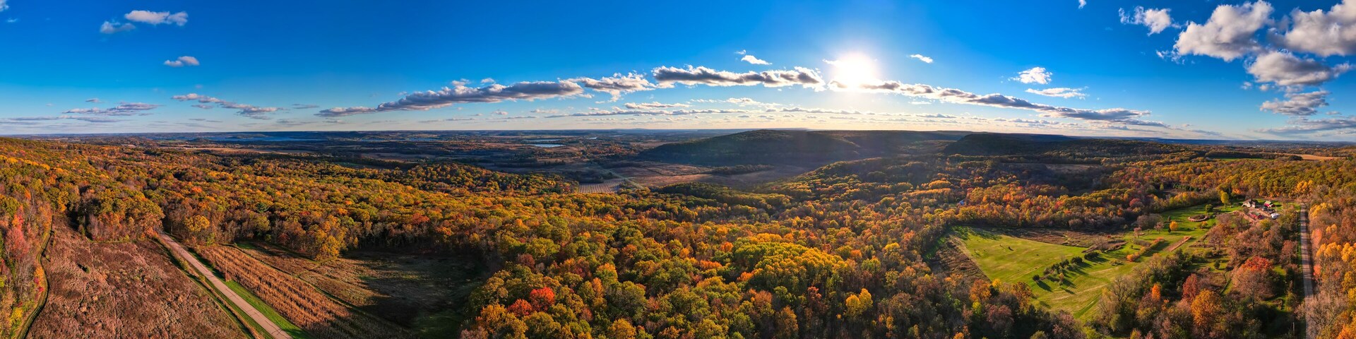 A panoramic view of a valley with autumn foliage, a winding road, and distant hills under a clear sky.
