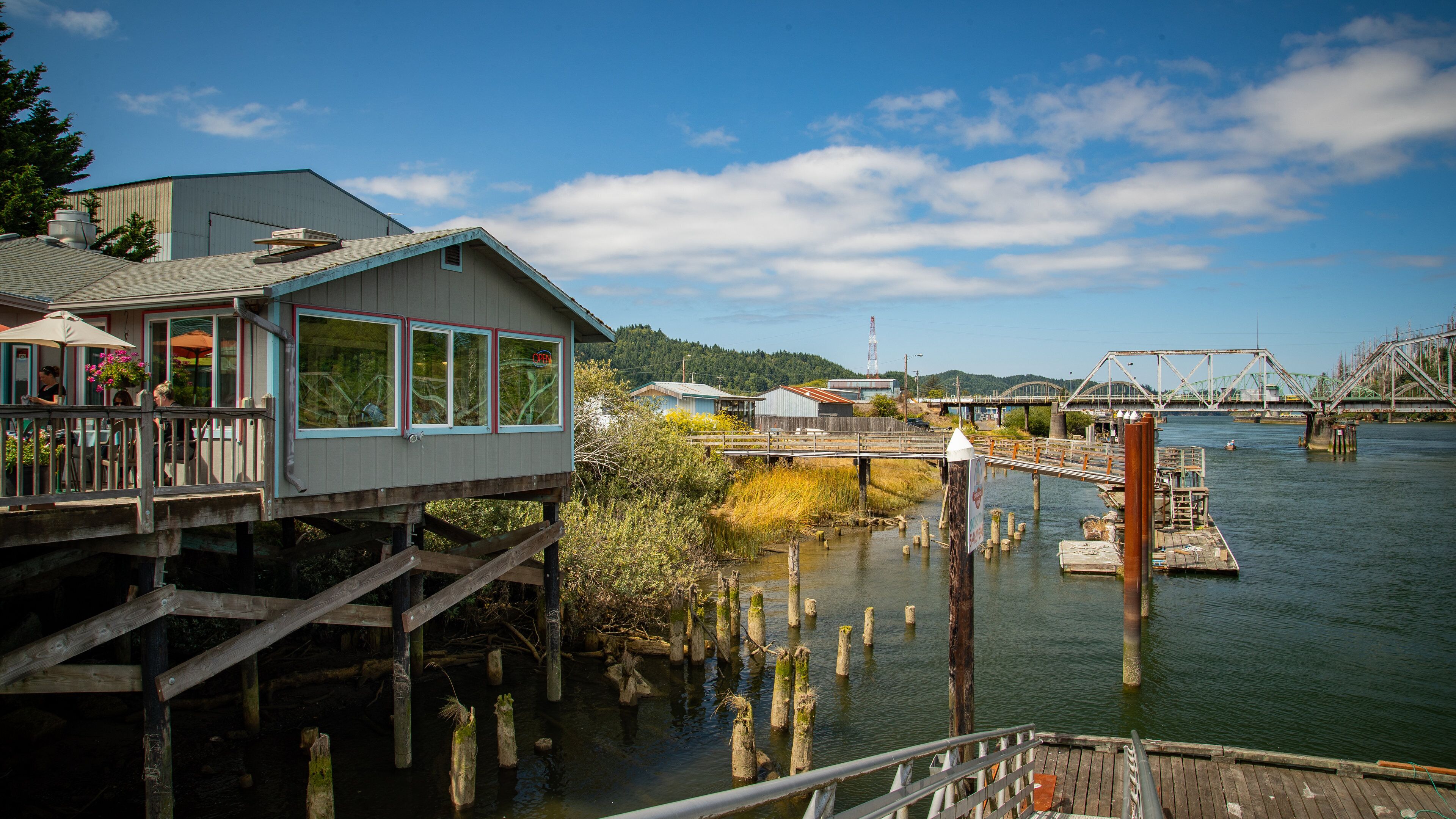 A riverside wooden deck with a house, a bridge, and industrial structures in the background.