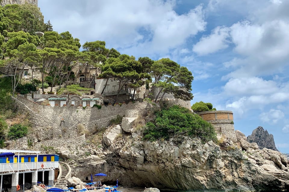Una zona costera con un acantilado rocoso, una playa con sombrillas azules y tumbonas, y una torre en el acantilado.