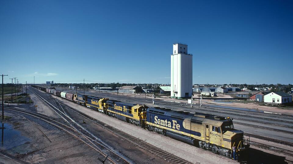 A train with 'Santa Fe' on the side moving along tracks in a rural area with a large white silo in the background.
