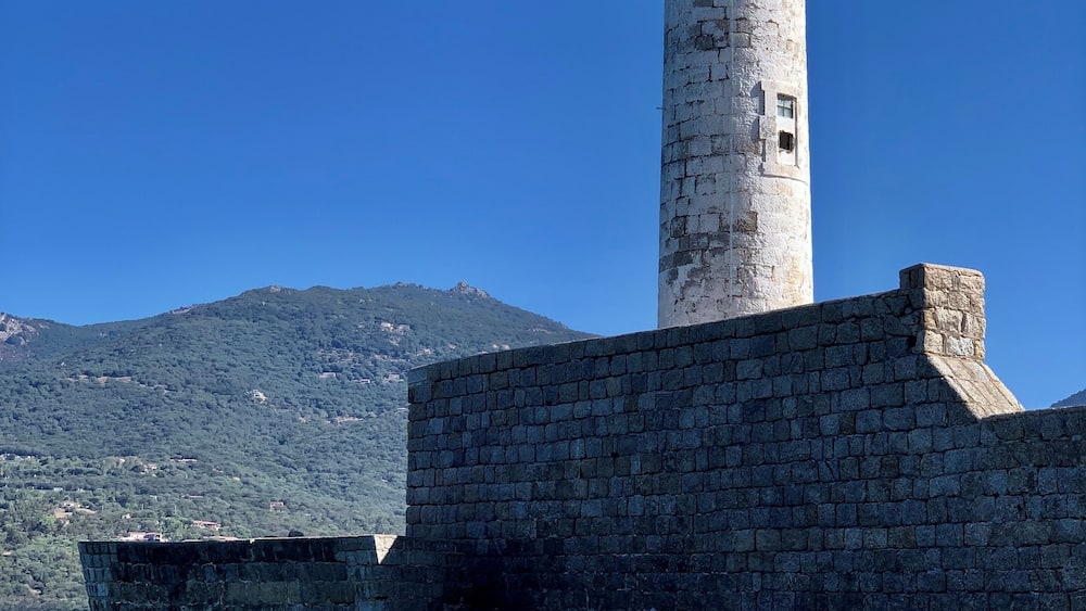 Un phare avec un sommet vert, un mur de pierre et une eau d’un bleu limpide.
