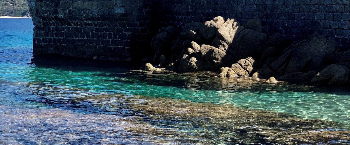A lighthouse with a green top, a stone wall, and clear blue water.