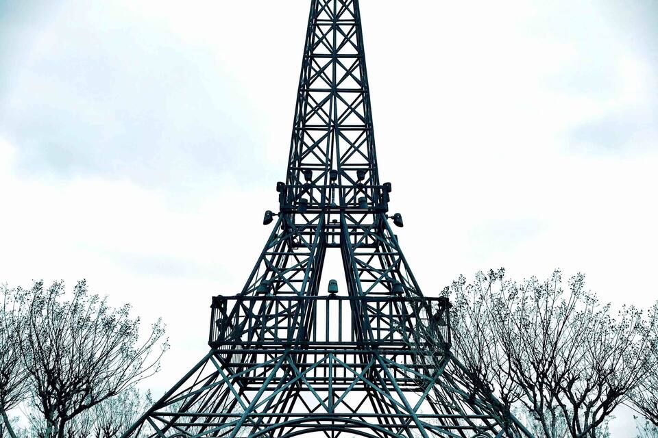 La Torre Eiffel con una antena parabólica grande en la parte superior, rodeada de árboles y una zona pavimentada.