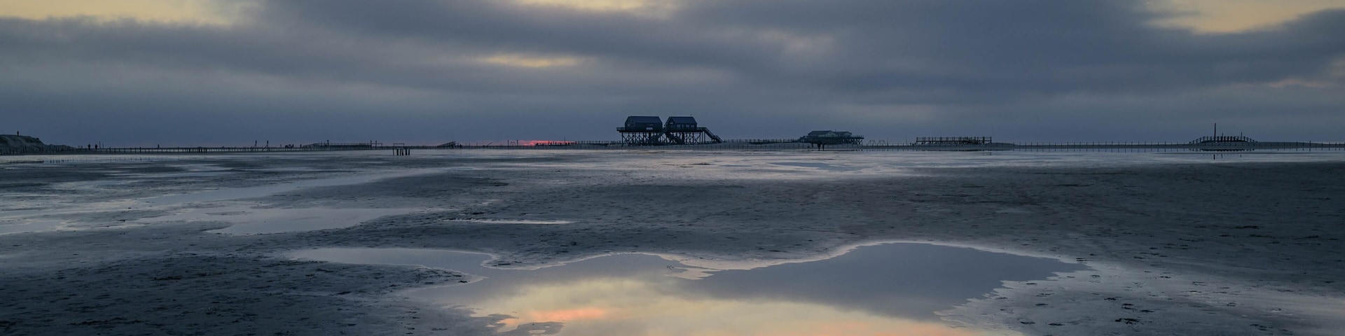 A coastal landscape with a pier extending into the water, a small structure on the pier, and a calm sea reflecting the sky.