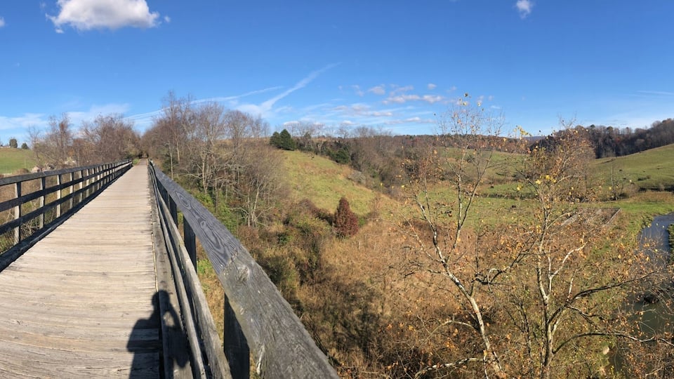 A wooden bridge over a river, surrounded by green hills and sparse trees.