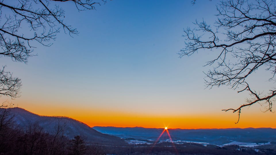 A sunset over a snow-covered landscape with bare trees and distant mountains.