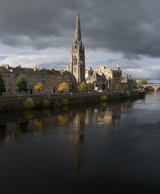 A river reflecting buildings, including a prominent tower, under a cloudy sky.