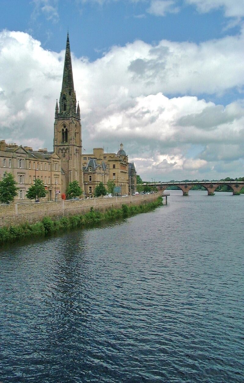 A historic church with a tall spire, a river, and a bridge in the background.