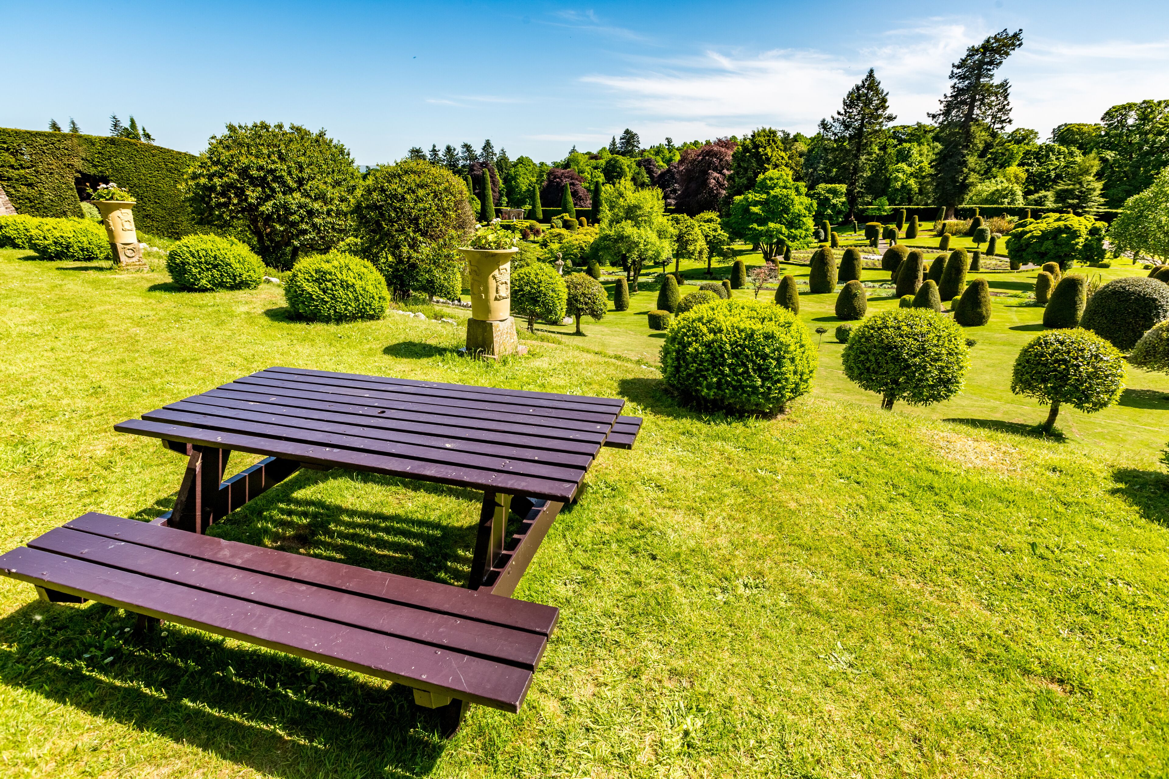 Mesa de piquenique num jardim bem cuidado, com sebes aparadas e diversas árvores.