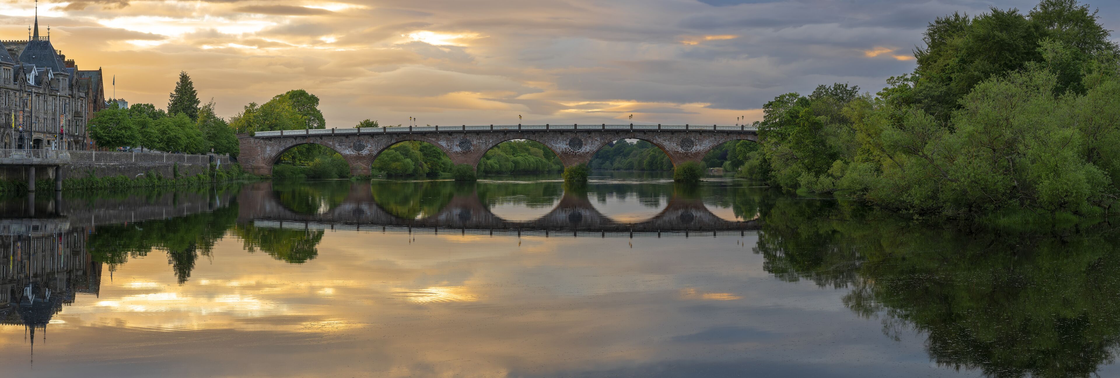 A historic stone bridge over a calm river, surrounded by lush greenery and a grand building with a spire.