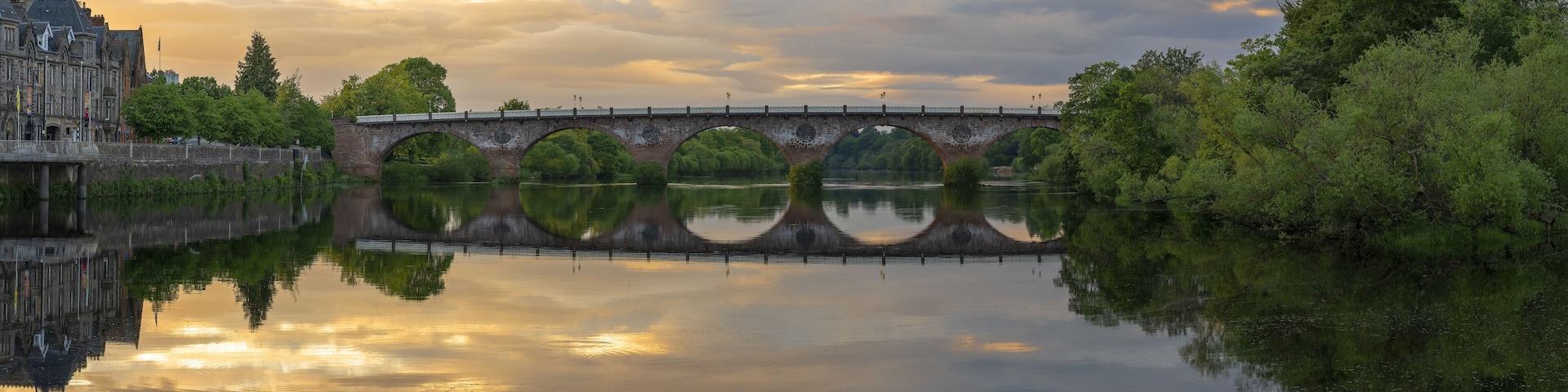 Un puente de piedra histórico sobre un río tranquilo, rodeado de exuberante vegetación y un imponente edificio con aguja.