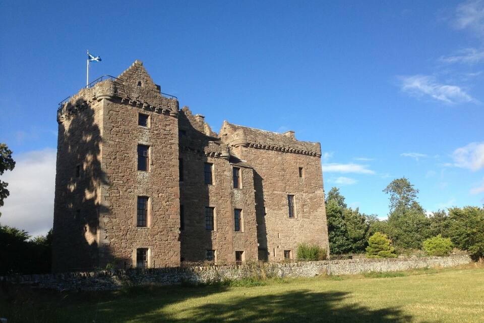 A historic stone castle with a flag on top, surrounded by greenery and trees.