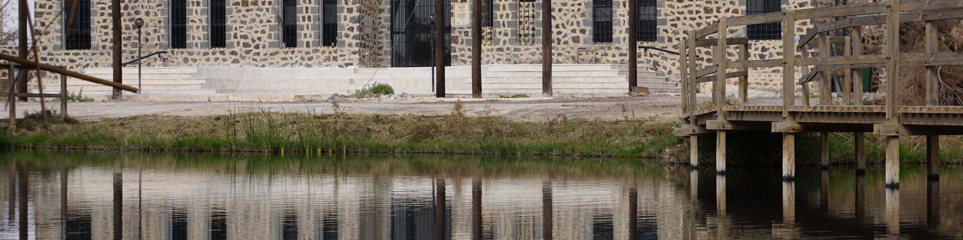 Ein Steinhaus mit Bogenfenstern und einer Holzbrücke über ruhigem Wasser.