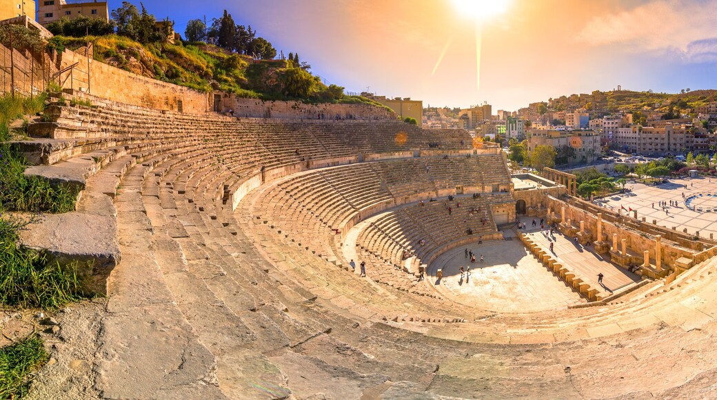 Un anfiteatro antiguo con escenario circular y asientos escalonados, rodeado por un paisaje urbano con edificios y colinas.