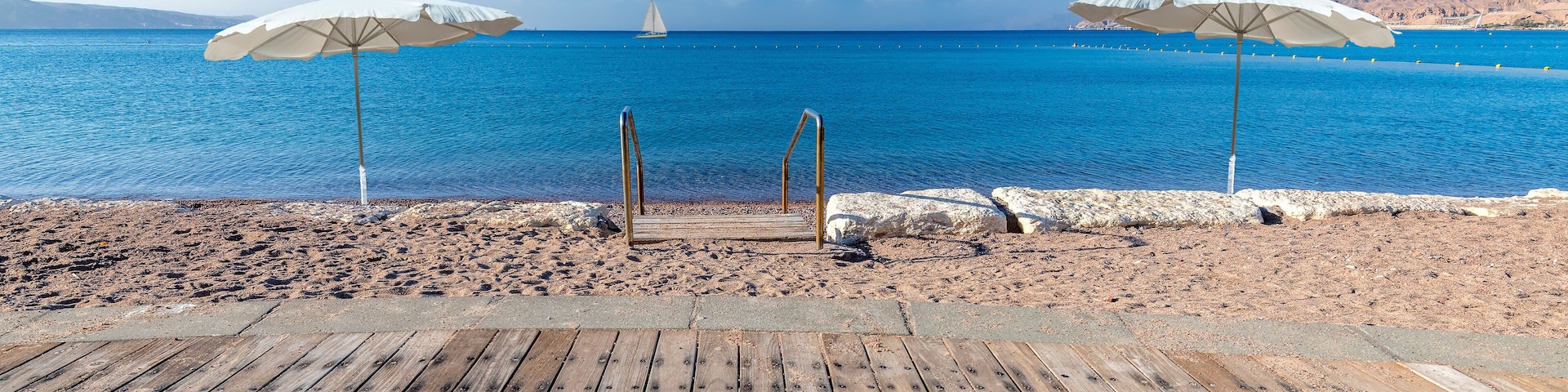 A wooden deck with two white beach umbrellas overlooking a beach with a sailboat in the distance.
