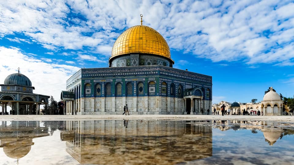 The Dome of the Rock with its golden dome and intricate blue and white mosaic patterns, reflecting in a puddle on the ground.