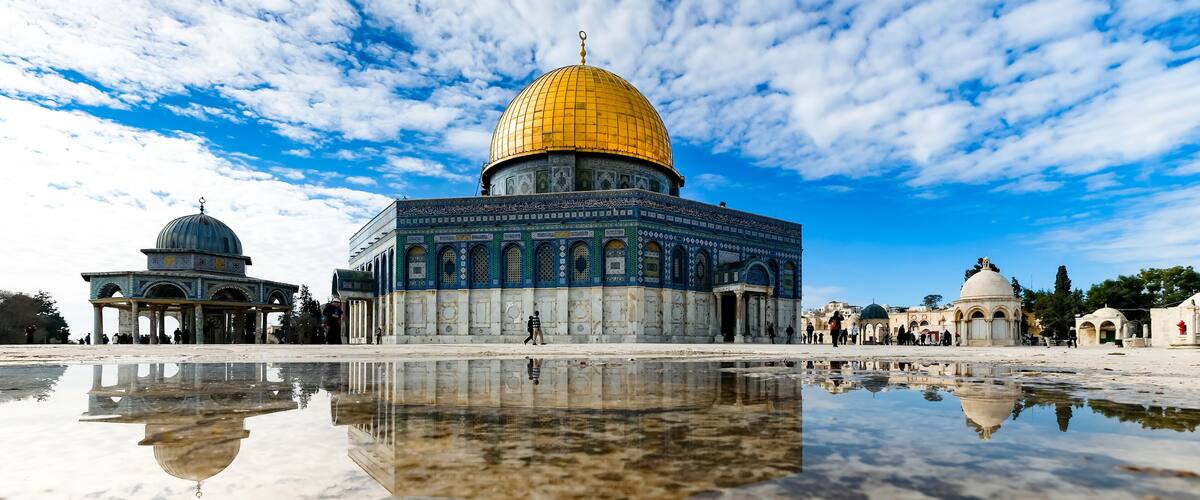 The Dome of the Rock with its golden dome and intricate blue and white mosaic patterns, reflecting in a puddle on the ground.