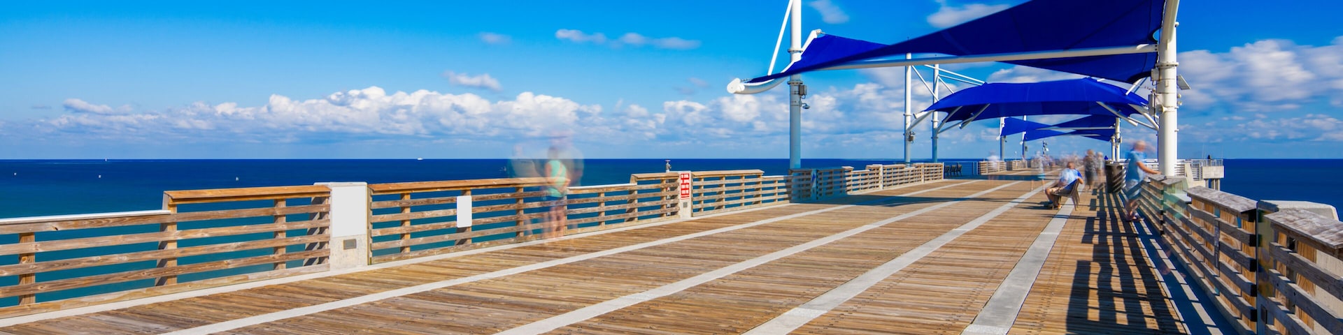 Una pasarela de madera con toldos azules, un cielo azul despejado y el océano al fondo.