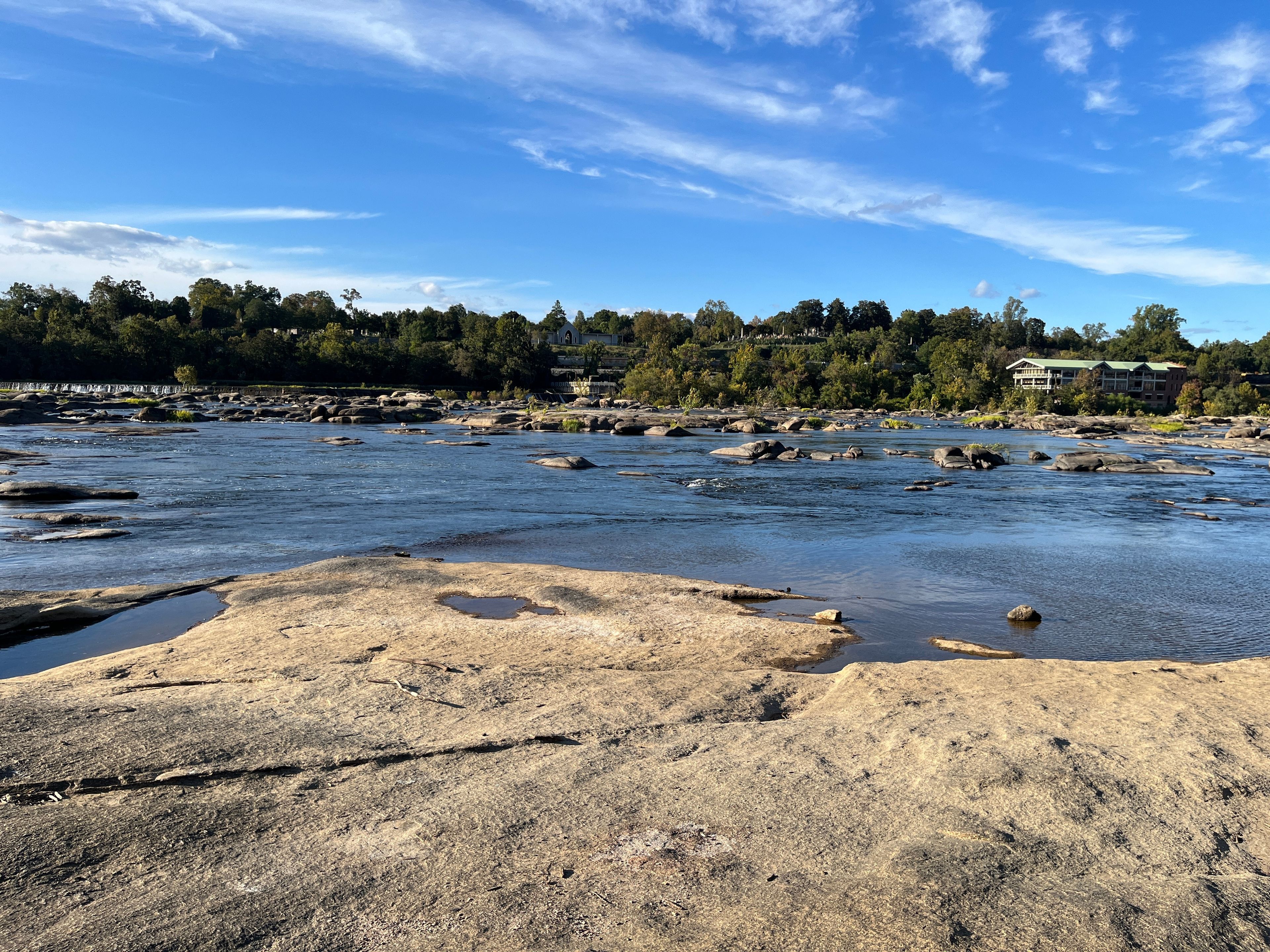 A river with rocks and a sandy foreground, a clear blue sky, and a forested hillside with buildings in the background.