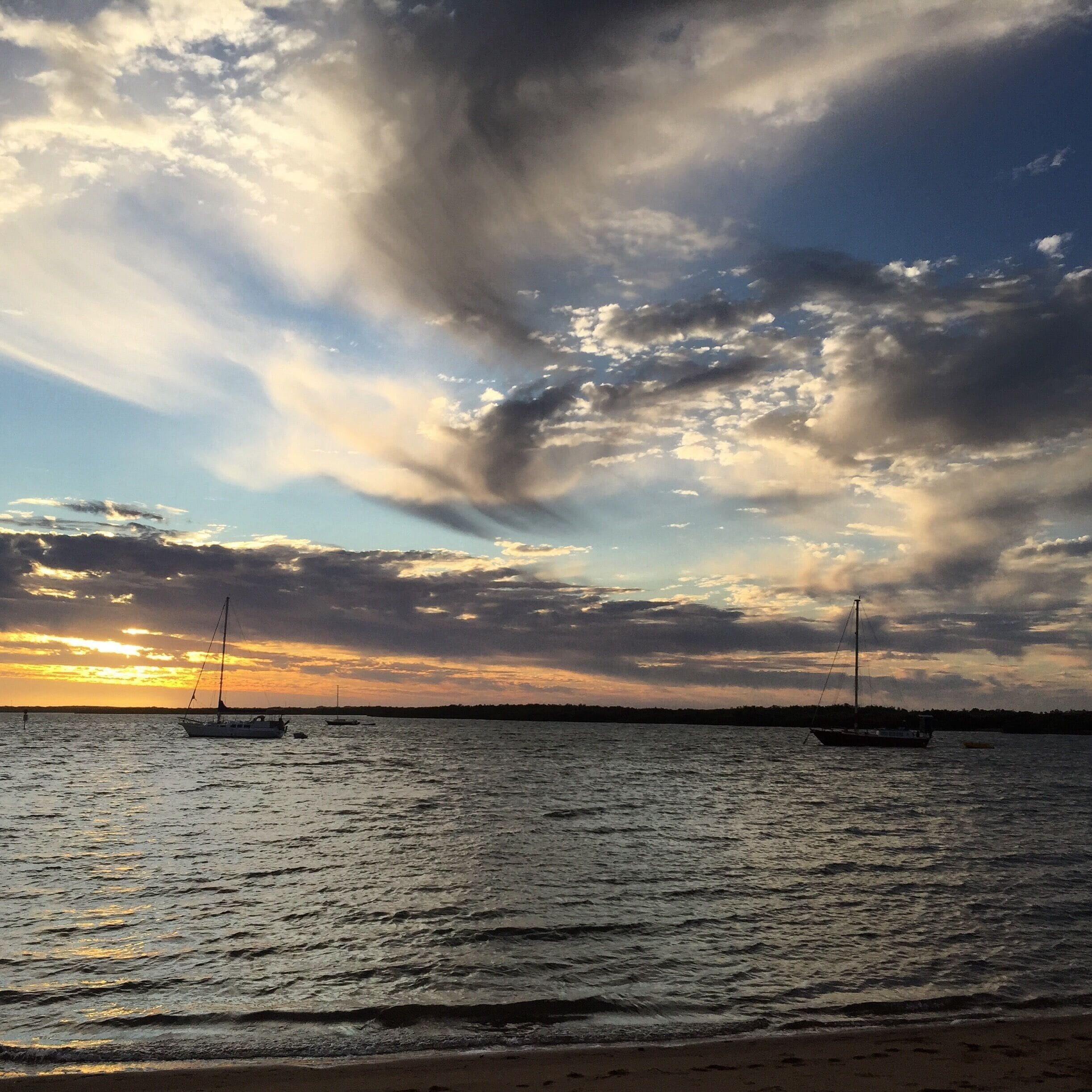 A sunset over a calm sea with sailboats and a beach.